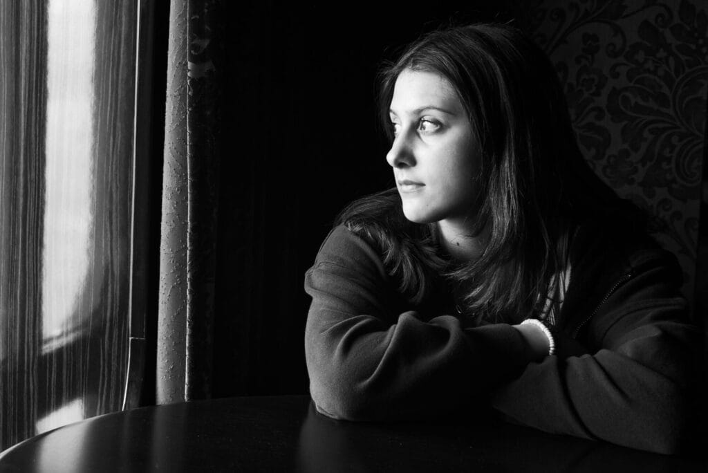 Black-and-white portrait of a young woman sitting beside a window, illuminated by natural light, photographed on traditional film.