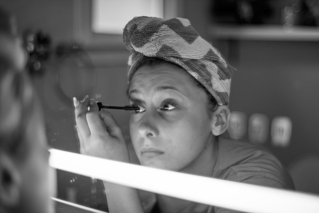 Black-and-white photograph of a young woman applying mascara in front of a mirror before a portrait session, captured on traditional film.