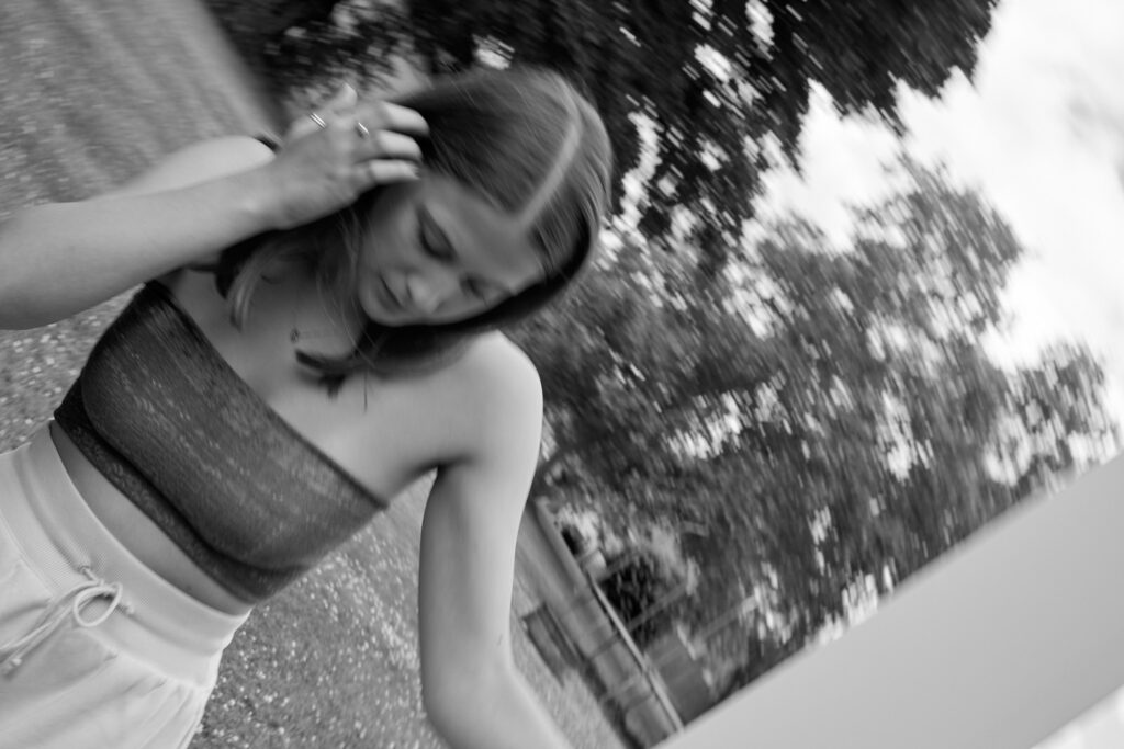 Black-and-white photo of a woman outdoors, reaching to touch her hair with one hand while the scene is motion-blurred and tilted.